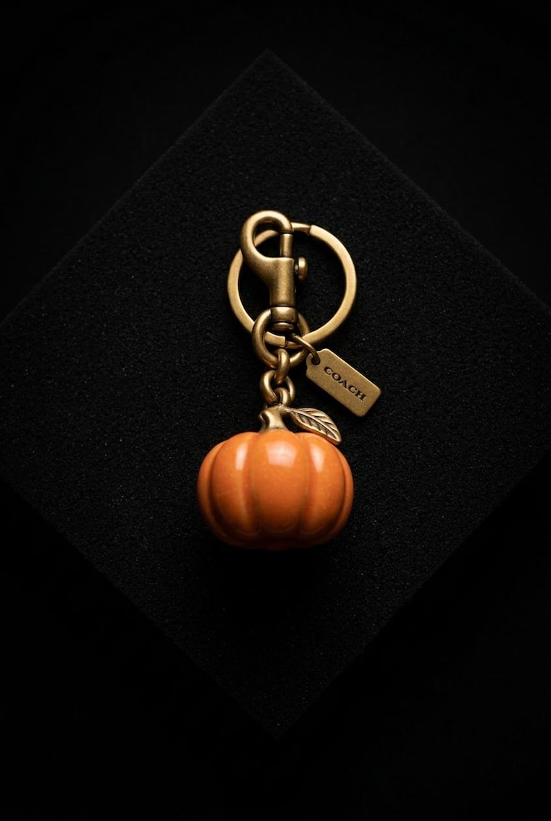 May include: A pumpkin-shaped charm keychain featuring a glossy orange pumpkin with a metallic stem and leaf. The charm is attached to a brass-toned metal split ring and a lobster clasp, accompanied by a small rectangular tag embossed with the word COACH.
