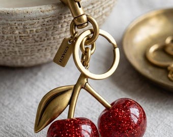 May include: A brass keychain featuring two round, glittery red cherry charms and a single brass leaf. The hardware includes a lobster clasp, a split ring, and a small rectangular tag embossed with the word COACH. The design is metallic and shiny.
