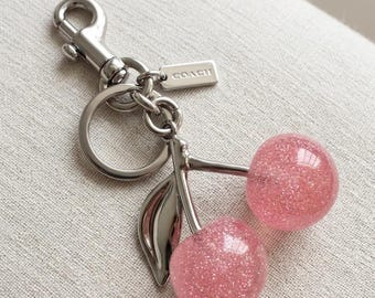 Könnte beinhalten: Ein Schlüsselanhänger aus silberfarbenem Metall mit zwei runden, mit rosa Glitzer gefüllten Kirschenanhängern, die an einem Schlüsselring und einem Karabinerhaken befestigt sind. Ein kleines rechteckiges Metallschild mit der Gravur COACH ist angebracht.