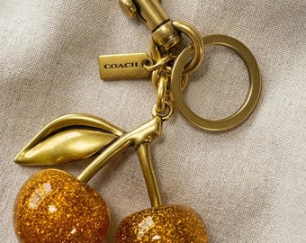 May include: A gold-toned metal keychain featuring two round, glittery amber-colored cherry charms attached to a stem with a single leaf. The hardware includes a lobster clasp, a circular split ring, and a small rectangular tag embossed with the word COACH.