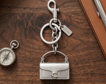 Könnte beinhalten: Ein silberfarbener Metall-Schlüsselanhänger mit einem Miniatur-Handtaschenanhänger, der einen Klappverschluss und ein kleines C-Logo aufweist. Der Schlüsselanhänger umfasst einen stabilen Karabinerhaken, einen Ring und ein kleines Schild mit Coach-Gravur.