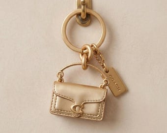 Könnte beinhalten: Ein goldfarbener Metallschlüsselanhänger mit einem Miniatur-Handtaschenanhänger, der einen Klappverschluss und ein markantes C-Logo aufweist. Der Anhänger ist an einem Ring befestigt und enthält ein kleines rechteckiges Schild mit dem Markennamen COACH.