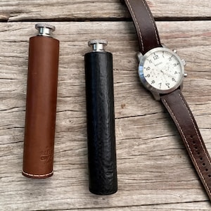 Peut inclure: Deux flasques enveloppées de cuir, une marron et une noire, avec des bouchons argentés. Une montre-bracelet avec un bracelet en cuir marron et un cadran blanc avec des chiffres noirs est également présente. Les articles sont disposés sur une surface en bois.