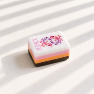 May include: A rectangular mahjong tile featuring a white top surface with a pink and purple koi fish graphic and the word JOKER printed in pink. The tile has a layered side profile consisting of a white, pink, orange, and dark brown base.