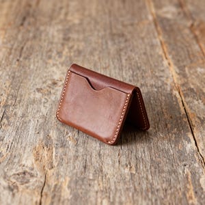 May include: A brown leather card holder with a minimalist design. The wallet features a single pocket on the front and is stitched with white thread. The leather has a smooth texture and a rich, warm color, set against a rustic wooden background.