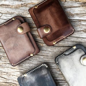 May include: Four leather cardholders in shades of brown, gray, and black are arranged on a wooden surface. Each cardholder features a snap closure with a brass button and white stitching. The leather has a distressed appearance.