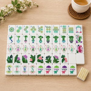 May include: A set of white rectangular mahjong tiles arranged in a grid, featuring colorful illustrations of plants, flowers, animals, and Chinese characters. The tiles include numbered suits, directional letters N, E, W, S, and Joker tiles with monkeys.