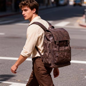 May include: A brown leather backpack with multiple pockets and a flap closure. The backpack has a distressed finish and adjustable straps. The person is wearing a tan shirt and brown pants.