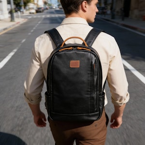May include: Black leather backpack with a brown leather handle and a small brown leather patch. The backpack has two side pockets and black shoulder straps. The zipper is black and the stitching is visible.