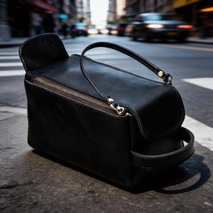 May include: A black leather bag with a rectangular body and a curved top flap. It features a gold-colored zipper and a small handle on top. The bag is sitting on a city street.