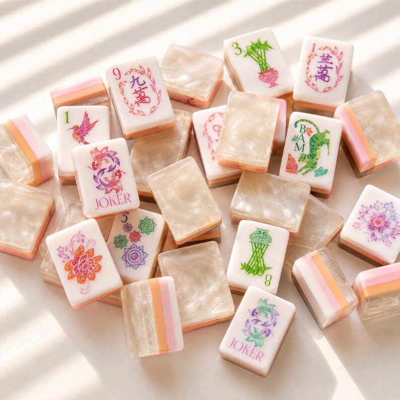 May include: A collection of mahjong tiles featuring a mix of white tiles with colourful illustrations of flowers, animals, and Chinese characters, alongside translucent, pearlescent beige tiles with pink and orange striped side accents.