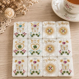 May include: Twelve rectangular white tiles with a pearlescent finish, arranged in a grid. Each tile features intricate gold embroidery, colorful rhinestones, and motifs including butterflies, hearts, clovers, crowns, and the word Joker.