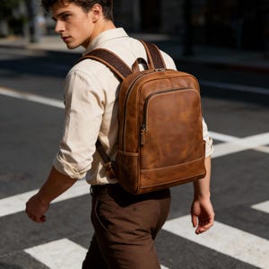 May include: A brown leather backpack with a rectangular shape and a front zippered pocket. The backpack has padded shoulder straps and a top handle. The stitching is a lighter brown color, and the hardware is silver. The person is walking on a crosswalk.