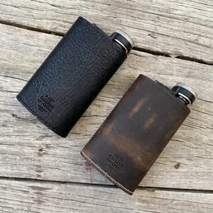 Könnte beinhalten: Zwei Lederflaschen, eine schwarze und eine braune, liegen auf einer Holzoberfläche. Die schwarze Flasche hat eine strukturierte Oberfläche, während die braune Flasche eine Used-Optik aufweist. Beide haben einen silbernen Verschluss und den Text "MADE BY DESERT GORGOO".