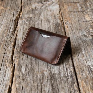 May include: A handcrafted, brown leather card holder with a minimalist design. The wallet features a single pocket and is stitched with light-colored thread. The leather has a rich, textured appearance, and the edges are neatly finished.