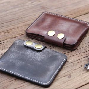 May include: Two leather wallets, one brown and one black, are displayed on a wooden surface. The brown wallet has a strap secured with two brass-colored buttons. The black wallet also has a strap with two brass-colored buttons. Both wallets are rectangular and have white stitching.