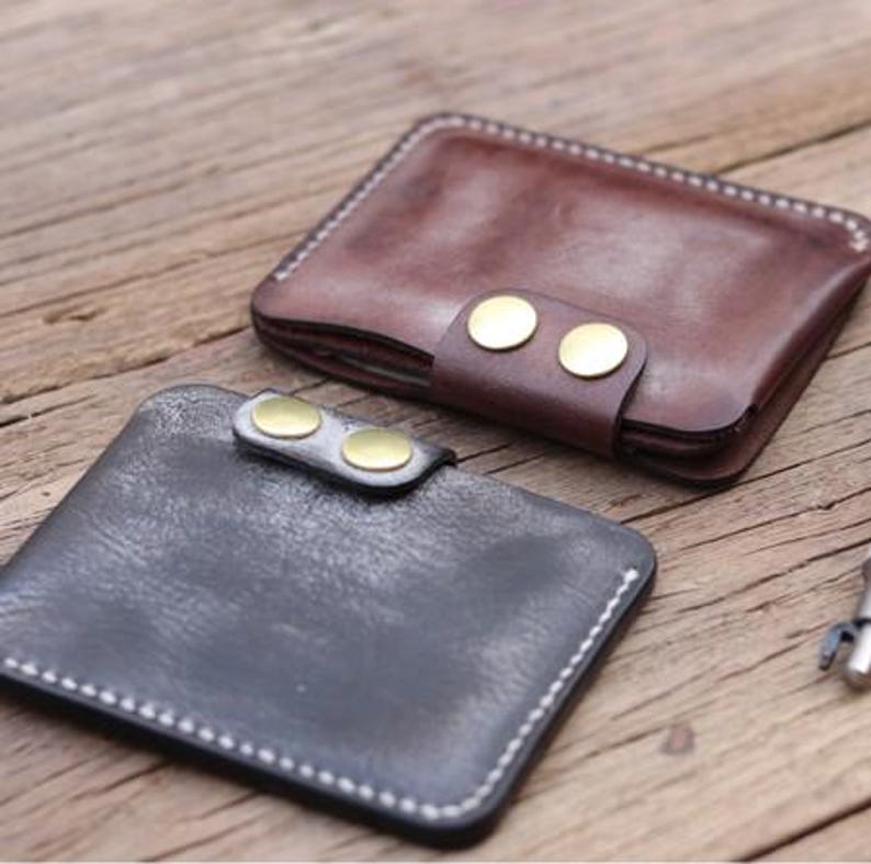 May include: Two leather wallets, one brown and one black, are displayed on a wooden surface. The brown wallet has a strap secured with two brass-colored buttons. The black wallet also has a strap with two brass-colored buttons. Both wallets are rectangular and have white stitching.