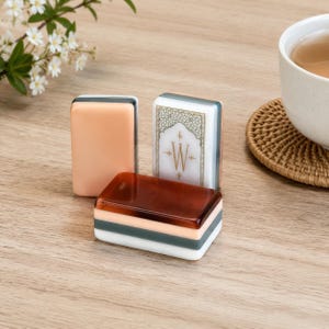 May include: Three rectangular mahjong tiles with smooth, glossy surfaces. One tile is solid peach, one is solid dark brown, and one is white with a gold-colored decorative border and the letter W centered within an intricate geometric pattern.