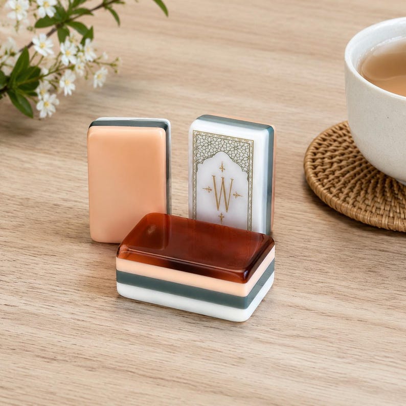 May include: Three rectangular mahjong tiles with smooth, glossy surfaces. One tile is solid peach, one is solid dark brown, and one is white with a gold-colored decorative border and the letter W centered within an intricate geometric pattern.