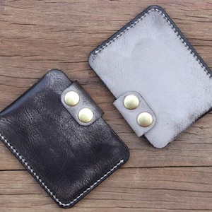 May include: Two leather cardholders in black and gray, each with a flap secured by two brass-colored snaps. The cardholders have white stitching around the edges and are displayed on a wooden surface.