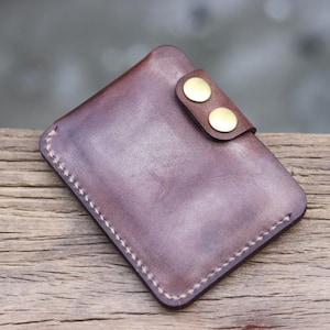 May include: A handcrafted, brown leather wallet with a snap closure. The wallet features a rectangular shape with rounded corners and visible white stitching around the edges. Two gold-colored snaps secure the flap.