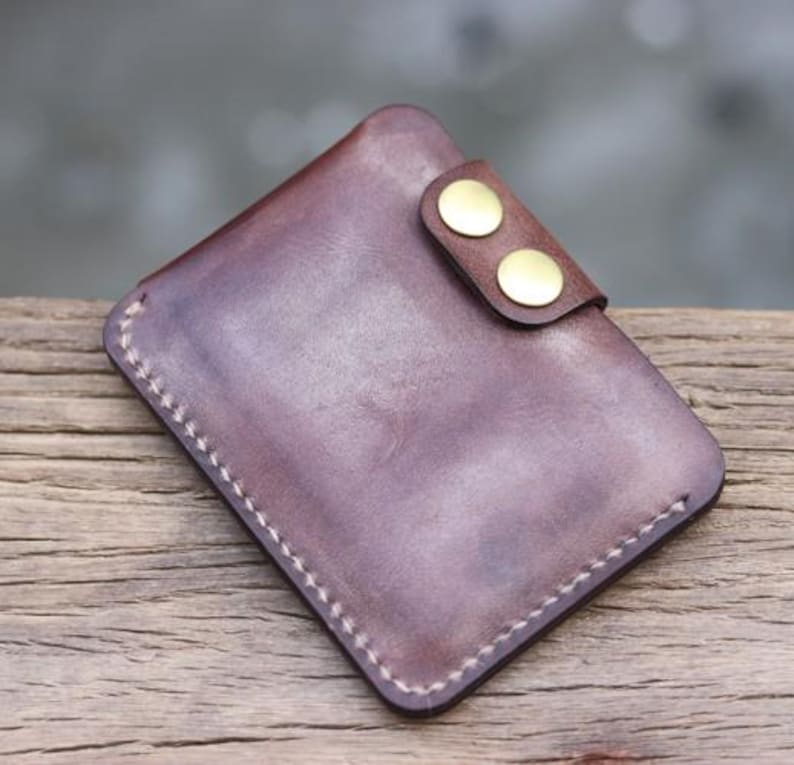 May include: A handcrafted, brown leather wallet with a snap closure. The wallet features a rectangular shape with rounded corners and visible white stitching around the edges. Two gold-colored snaps secure the flap.