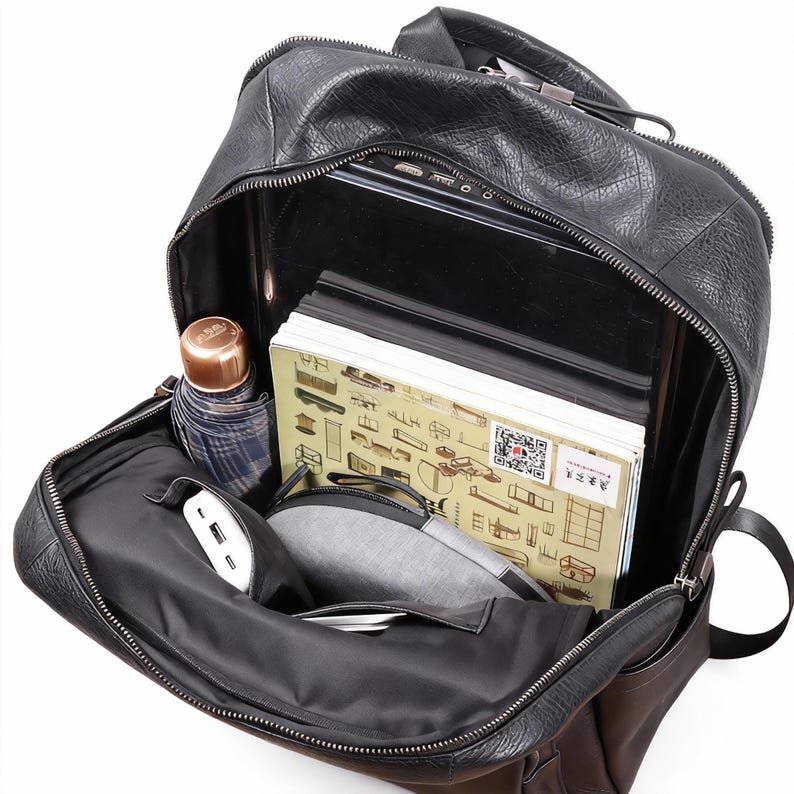 Op de afbeelding: Zwarte leren rugzak met een open rits, met daarin een laptop, boeken, een kleine paraplu en een draagbare oplader. De tas heeft meerdere compartimenten en een handvat aan de bovenkant. De binnenkant is van zwarte stof.