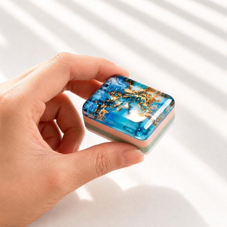 May include: A small square container with a glossy top featuring a marbled blue and gold pattern. The sides of the container are layered with bands of light pink, light green, and white, creating a compact and smooth rectangular object.