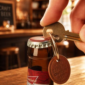 Op de afbeelding: Een bruine bierfles met een zilveren en rood etiket wordt geopend met een sleutel. Een leren label met de tekst "MAKE LIFE DESSERT City of God SINCE 2003" hangt aan de sleutelhanger. De achtergrond is een bar.