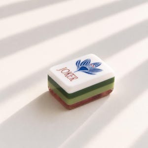 May include: A rectangular mahjong tile featuring a white top surface with the word JOKER printed in brown and a blue floral graphic. The tile has a layered side profile consisting of a white top layer, a light green middle layer, and a dark green base.