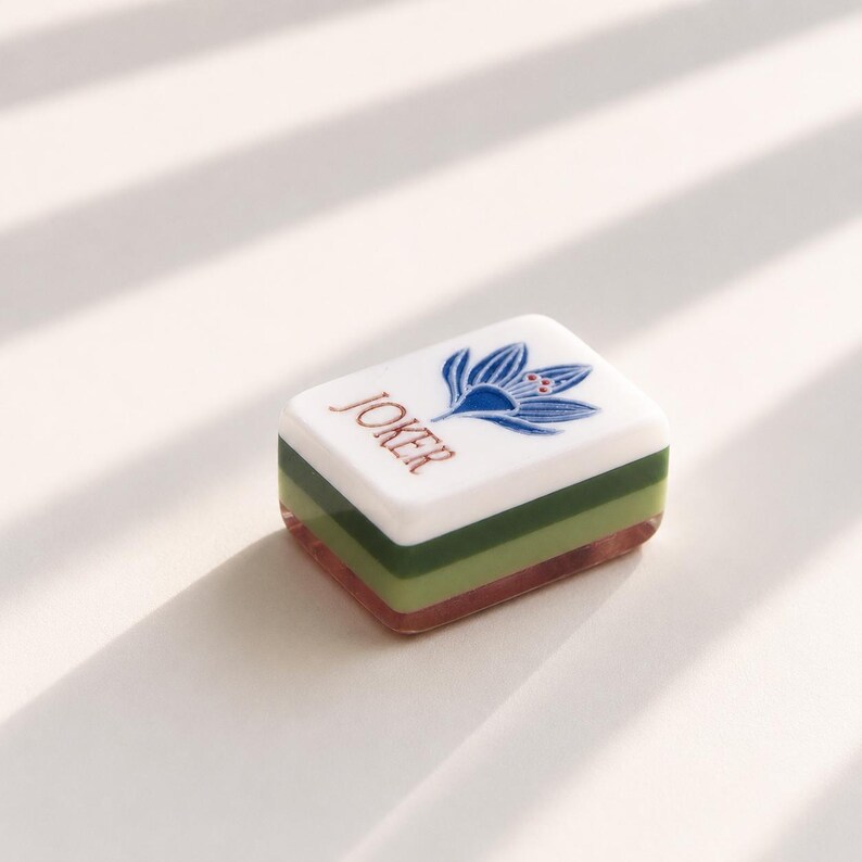 May include: A rectangular mahjong tile featuring a white top surface with the word JOKER printed in brown and a blue floral graphic. The tile has a layered side profile consisting of a white top layer, a light green middle layer, and a dark green base.