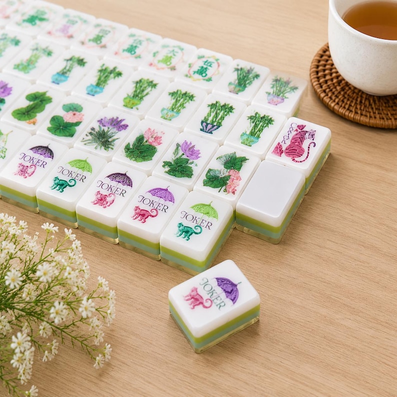 May include: A set of rectangular mahjong tiles with white tops and layered green bases. The tiles feature colorful illustrations of plants, flowers, and animals. Several tiles are labeled with the word JOKER beneath a purple or green umbrella.