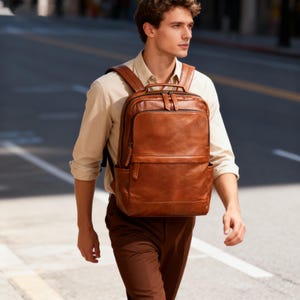 Puede incluir: Una mochila de cuero marrón con múltiples compartimentos y un asa superior. La mochila tiene forma rectangular con un bolsillo frontal y bolsillos laterales. El cuero tiene un aspecto texturizado y un color rico y cálido.