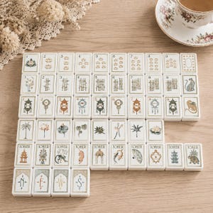 May include: A set of rectangular mahjong tiles arranged in rows, featuring white surfaces with gold-toned borders. The tiles display various illustrations including numbers one through nine, floral motifs, bamboo, animals, lanterns, and joker cards with text.