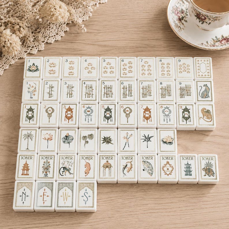 May include: A set of rectangular mahjong tiles arranged in rows, featuring white surfaces with gold-toned borders. The tiles display various illustrations including numbers one through nine, floral motifs, bamboo, animals, lanterns, and joker cards with text.
