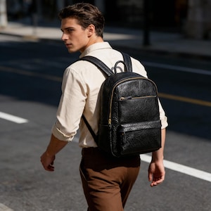 May include: Black textured leather backpack with gold-colored zippers and a top handle. The backpack has a front pocket and adjustable shoulder straps. The bag is being worn by a person walking on a city street.