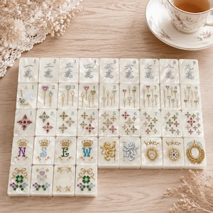 May include: A set of white pearlescent mahjong tiles arranged in a grid. The tiles feature engraved numbers one through nine, colorful gemstone-style star and wand motifs, letters N, S, E, W, lion figures, joker text, and floral clover designs.