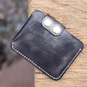 May include: A small, black leather wallet with a textured surface. The wallet has a flap secured by two gold-colored button snaps. White stitching outlines the edges of the wallet, which appears to be handmade.
