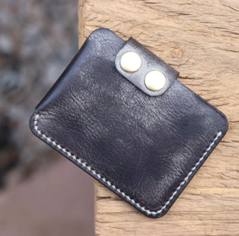 May include: A small, black leather wallet with a textured surface. The wallet has a flap secured by two gold-colored button snaps. White stitching outlines the edges of the wallet, which appears to be handmade.