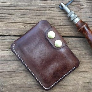May include: A brown leather wallet with a snap closure and two brass-coloured buttons. The wallet has white stitching around the edges and is set on a wooden surface. A leatherworking tool is visible in the background.