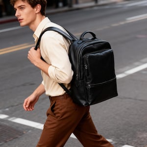 Puede incluir: Una mochila de cuero negro con múltiples compartimentos con cremallera y un asa superior. La mochila tiene correas acolchadas para los hombros y un bolsillo frontal rectangular. La persona de la imagen lleva una camisa clara y pantalones marrones.