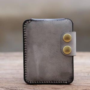 May include: A gray leather card holder with a distressed finish. The wallet features a flap secured by two brass-colored snap closures. The edges are stitched with a contrasting thread, and the overall design is minimalist and functional.