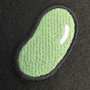 K&ouml;nnte beinhalten: Ein gestickter Aufn&auml;her in Form einer Kidneybohne. Der Aufn&auml;her hat eine hellgr&uuml;ne Mitte mit einem kleinen wei&szlig;en Glanzpunkt auf der rechten Seite, eingefasst von einem dicken, schwarz gestickten Rand, der die geschwungene ovale Form betont.