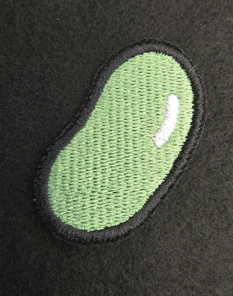 K&ouml;nnte beinhalten: Ein gestickter Aufn&auml;her in Form einer Kidneybohne. Der Aufn&auml;her hat eine hellgr&uuml;ne Mitte mit einem kleinen wei&szlig;en Glanzpunkt auf der rechten Seite, eingefasst von einem dicken, schwarz gestickten Rand, der die geschwungene ovale Form betont.