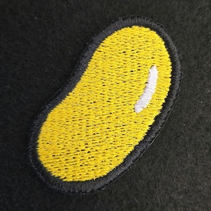 K&ouml;nnte beinhalten: Ein gestickter Aufn&auml;her in Form einer gelben Geleebohne mit schwarzem Rand und einer kleinen wei&szlig;en Markierung auf der oberen rechten Seite. Die Stickerei besteht aus dichtem gelbem Garn, das die Bohnenform mit einem schwarzen Rand f&uuml;llt.