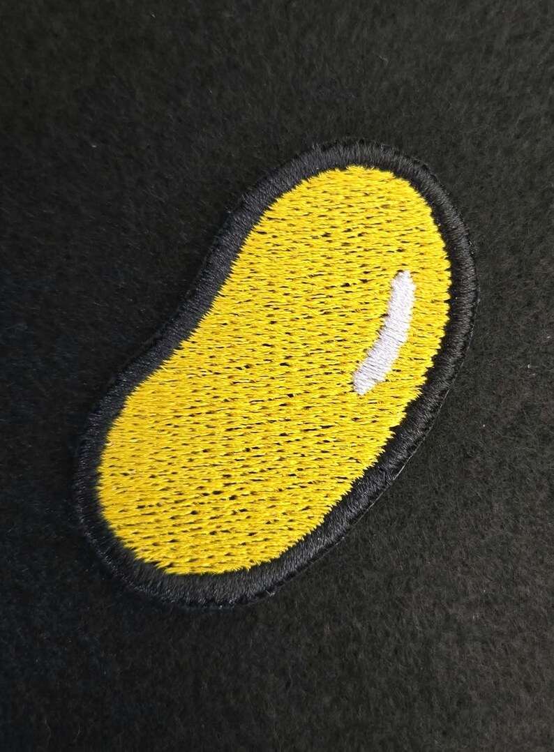 K&ouml;nnte beinhalten: Ein gestickter Aufn&auml;her in Form einer gelben Geleebohne mit schwarzem Rand und einer kleinen wei&szlig;en Markierung auf der oberen rechten Seite. Die Stickerei besteht aus dichtem gelbem Garn, das die Bohnenform mit einem schwarzen Rand f&uuml;llt.