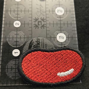 K&ouml;nnte beinhalten: Ein ovaler, roter Stickaufn&auml;her mit schwarzem Rand und einem kleinen wei&szlig;en, halbmondf&ouml;rmigen Akzent. Der Aufn&auml;her liegt auf einem transparenten Kunststofflineal, das eine Breite von ca. 3,8 cm und eine H&ouml;he von ca. 1,9 cm anzeigt.