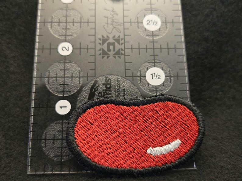 K&ouml;nnte beinhalten: Ein ovaler, roter Stickaufn&auml;her mit schwarzem Rand und einem kleinen wei&szlig;en, halbmondf&ouml;rmigen Akzent. Der Aufn&auml;her liegt auf einem transparenten Kunststofflineal, das eine Breite von ca. 3,8 cm und eine H&ouml;he von ca. 1,9 cm anzeigt.