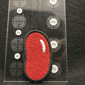 K&ouml;nnte beinhalten: Ein roter, nierenf&ouml;rmiger Stickaufn&auml;her mit schwarzem Rand und einem kleinen wei&szlig;en Glanzlicht, platziert neben einem transparenten Kunststofflineal, das zeigt, dass der Aufn&auml;her etwa 3,8 cm hoch und 1,9 cm breit ist.