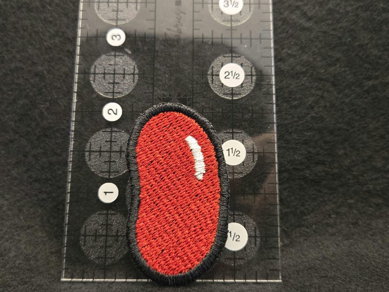 K&ouml;nnte beinhalten: Ein roter, nierenf&ouml;rmiger Stickaufn&auml;her mit schwarzem Rand und einem kleinen wei&szlig;en Glanzlicht, platziert neben einem transparenten Kunststofflineal, das zeigt, dass der Aufn&auml;her etwa 3,8 cm hoch und 1,9 cm breit ist.